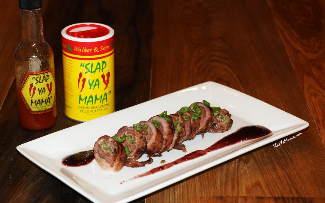 Stuffed Duck Breasts Wrapped In Bacon with a Black Cherry Sauce on a rectangular white plate with the seasoning positioned behind and to the left.