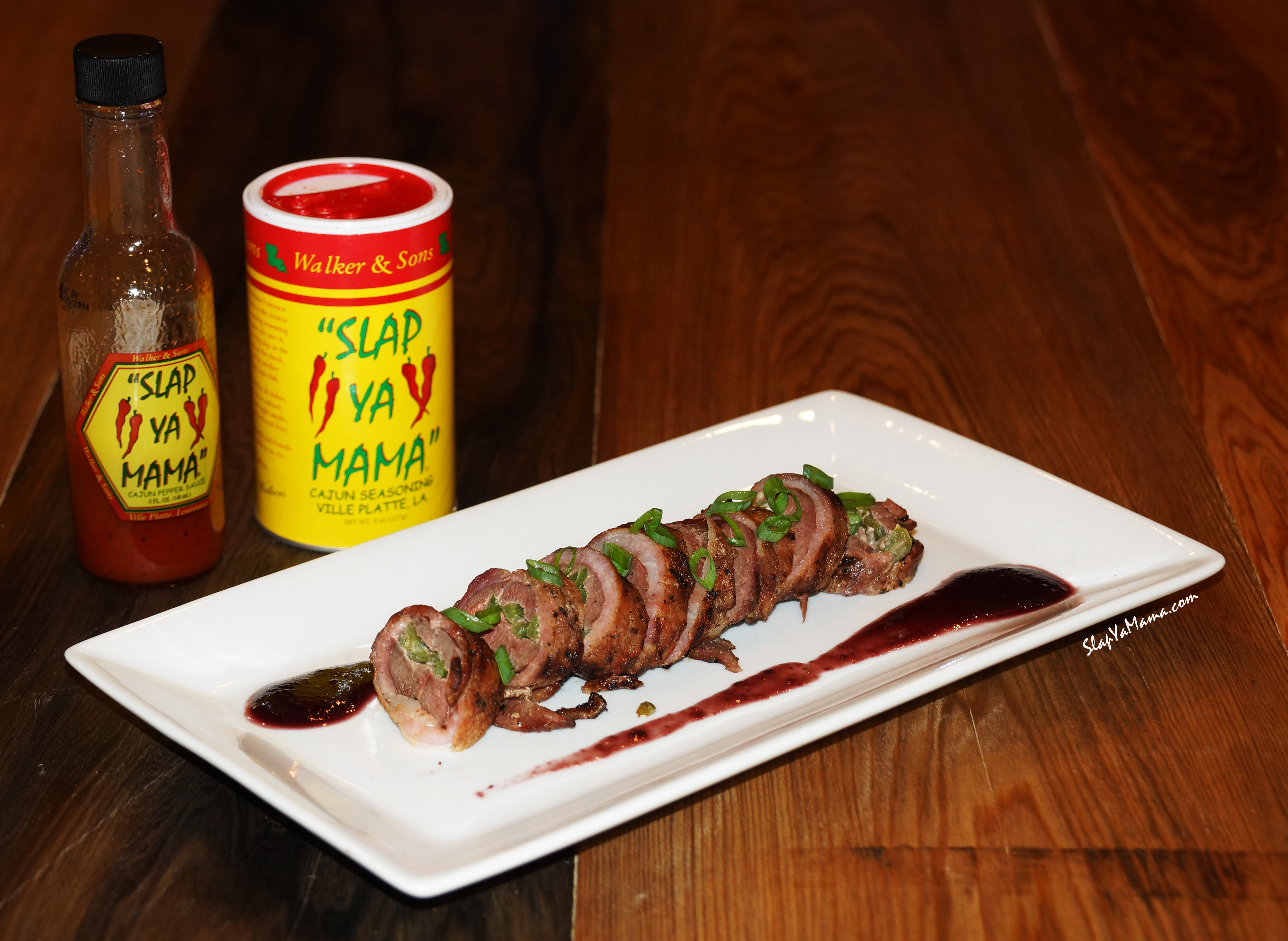 Stuffed Duck Breasts Wrapped In Bacon with a Black Cherry Sauce on a rectangular white plate with the seasoning positioned behind and to the left.